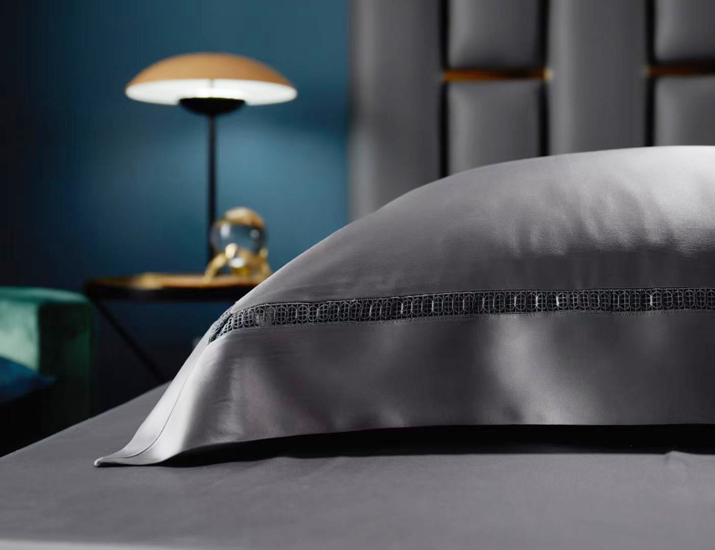 A close-up view of a gray pillow with a decorative trim, placed on a bed with a dark blue wall in the background.