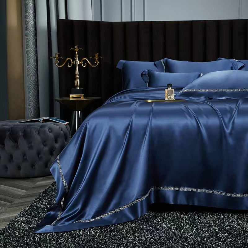 A luxurious bedroom with a large bed covered in a blue satin comforter and matching pillows. The bed is framed by a dark headboard, and a gold candelabra is placed on the nightstand next to the bed. The room has a plush gray rug and a black tufted ottoman, creating a sophisticated and elegant atmosphere.