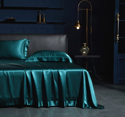 A luxurious bedroom with a dark blue color scheme, featuring a bed with a satin bedspread and matching pillows, a nightstand with a lamp, and a gold-colored lampshade.
