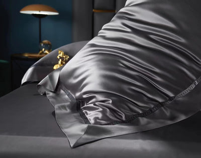 A close-up view of a luxurious gray satin bedding set, including a pillow and a blanket, with a gold-colored decorative pillow in the background.