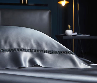 A close-up view of a gray silk pillowcase with a decorative silver trim, resting on a bed.