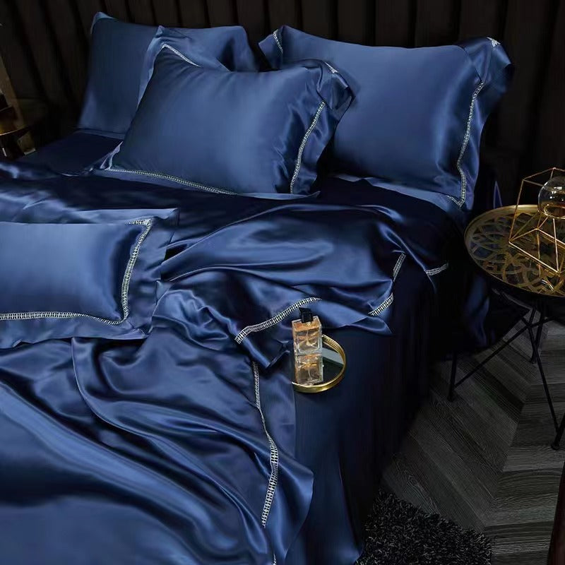 A luxurious bed with a blue satin comforter and matching pillows, complemented by a gold-trimmed nightstand with a glass container.